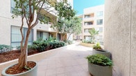 Located at 606 3rd Street in San Diego, this outdoor courtyard serves as the central communal space of Market Street Square Apartment Homes. The courtyard is defined by its open-air design, framed on multiple sides by the multi-story residential buildings whose warm terracotta and earth-toned stucco facades rise above the space, providing an enclosed yet airy atmosphere.

The ground level is finished with light-toned, grid-patterned tile pavers that form wide, accessible walkways throughout the courtyard. Generous landscaping beds line the perimeter and interior of the space, planted with a variety of shrubs, ornamental grasses, tropical foliage, and mature shade trees whose canopies extend overhead, providing natural coverage and dappled light. Large, round concrete planters are thoughtfully distributed along the walkways, adding further greenery and visual interest.

A fenced water feature with a fountain serves as a focal point toward one end of the courtyard, contributing both visual appeal and ambient sound. The textured stucco walls of the buildings catch natural sunlight throughout the day, creating dynamic light and shadow patterns across the space. Building access is provided through glass-paneled entry doors set into the stucco facade. Overall, the courtyard offers a calm, well-maintained outdoor environment within an urban San Diego setting.