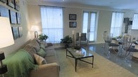 This single-floor space centers around a well-appointed living room that balances comfort and functionality. Large windows fitted with white horizontal blinds allow generous natural light to fill the room, creating a bright and airy atmosphere. A set of glass-paned French doors further enhances the sense of openness, providing an additional source of light. The flooring features dark hardwood, which contrasts elegantly with a woven natural fiber area rug that anchors the seating arrangement. The main seating area includes a spacious, neutral-toned sectional sofa adorned with decorative green and tan throw pillows, complemented by a patterned accent chair in blue and green hues. A sleek glass-top coffee table with a dark metal frame serves as the focal point of the arrangement, topped with decorative books and accessories. Tall pillar candles on slender stands add a subtle decorative touch. Lush indoor plants positioned near the windows bring a natural, organic element into the space. Wall décor includes two framed prints, adding a personalized character to the neutral walls. A floor lamp in the corner provides warm ambient lighting, contributing to the overall inviting and relaxed ambiance of the living area.