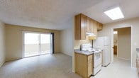 This single-floor residence offers an open and functional layout that seamlessly connects the kitchen and living areas. The kitchen is equipped with white appliances, including an electric range and a full-sized refrigerator, complemented by warm-toned wood cabinetry that provides ample storage above and below the counter. A granite-style countertop adds a practical workspace, while a built-in range hood sits beneath the upper cabinets. A skylight overhead brings in additional natural light, brightening the kitchen area. The living space features neutral wall-to-wall carpeting and beige walls, contributing to a clean, understated aesthetic. A textured popcorn ceiling runs throughout, consistent with the overall design of the unit. A large sliding glass door along the far wall of the living area floods the space with natural light and provides direct access to an outdoor balcony, offering a seamless connection between interior and exterior. The overall ambiance is practical and straightforward, with a layout that prioritizes functionality and ease of movement throughout the open floor plan.