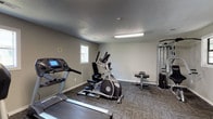 This fitness center is a purpose-built, single-floor space designed to support a well-rounded workout routine. The room features neutral gray walls, textured ceilings, and durable patterned carpet tiles throughout, creating a clean and functional environment. Natural light filters in through multiple windows positioned along the walls, complemented by overhead fluorescent lighting to ensure the space remains bright and well-lit at all times.

The cardio section is well-stocked, offering multiple treadmills, an elliptical trainer, and a stationary recumbent bike, providing a range of options for low- and high-impact cardiovascular exercise. For strength training, the space includes a multi-station cable machine capable of supporting a variety of resistance exercises, along with a weight bench, free weights, and additional resistance equipment. A dedicated entry door with a glass panel provides convenient access to the space, and a stainless steel waste bin is thoughtfully placed near the equipment area for easy upkeep. A small wall-mounted shelf offers a convenient spot for personal items during workouts. The layout is open and spacious, allowing users to move freely between equipment without feeling crowded, making this fitness center a practical and accessible amenity.