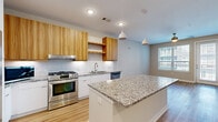 This single-floor unit showcases a thoughtfully designed open layout with light hardwood flooring flowing seamlessly throughout every room. The kitchen serves as the centerpiece, featuring a large central island topped with granite countertops and pendant lighting overhead. Warm wood-toned upper cabinetry contrasts stylishly with crisp white lower cabinets and a classic subway tile backsplash. Stainless steel appliances, including a gas range, dishwasher, and a French-door refrigerator, complete the well-equipped space.

The living and dining areas are bright and open, benefiting from multiple windows fitted with horizontal blinds that allow natural light to filter in. Several rooms include access to private exterior balconies through glass-panel doors, offering outdoor connectivity. Ceiling fans are installed in key living spaces, enhancing comfort and airflow.

A dedicated laundry area houses a stacked washer and dryer unit, conveniently positioned within the floor plan. The bathroom features accessible design elements, including grab bars and hardwood-style flooring consistent with the rest of the unit. Neutral wall tones throughout create a clean, cohesive aesthetic that complements the modern finishes.