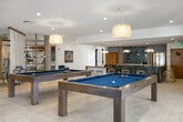 Located at 1515 2nd Street in Coronado, California, this generously proportioned single-floor space offers a thoughtfully designed layout ideal for gathering and recreation. The interior showcases a coastal-modern aesthetic, with light-toned tile flooring, recessed lighting, and an open, airy atmosphere throughout.

Two distinct living areas provide flexible seating arrangements, each characterized by elegant textile room dividers that add an artistic, bohemian touch while maintaining a sense of flow between spaces. Natural fiber rugs, leather and upholstered furnishings, and curated artwork contribute to a warm yet contemporary ambiance.

The dining room features a large white-topped communal table surrounded by woven rattan chairs, anchored by a sculptural woven pendant light. Floor-to-ceiling glass doors invite natural light and offer views of lush outdoor greenery, seamlessly connecting the interior to the outdoors.

The expansive game room is a standout feature, housing two full-size pool tables beneath pendant fixtures, with large black-framed windows framing views of surrounding tropical landscaping. An additional media room offers a recessed flat-screen display and a bold geometric accent wall. Open-plan social areas with bar-height seating and wood shelving complete this versatile and inviting space.