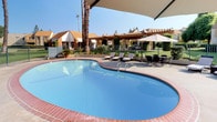 This inviting outdoor amenity space centers around a generously sized freeform swimming pool with striking turquoise water and classic brick coping that adds a timeless aesthetic touch. The pool deck is designed for relaxation and leisure, outfitted with multiple cushioned chaise lounges and rattan-style seating arranged to take full advantage of the open-air environment. Large cantilever umbrellas provide ample shade over key seating areas, making the space comfortable even under bright, sunny skies. A dedicated dining or lounge grouping with a table and chairs offers an area for casual outdoor entertaining. The surrounding landscape is lush and well-maintained, featuring manicured shrubs, flowering plants, and mature trees that create a sense of privacy and a tranquil garden atmosphere. A wrought iron perimeter fence encloses the area, offering security while maintaining sightlines to the verdant greenery beyond. A pergola structure is visible in the background, adding an architectural layer to the outdoor setting. The warm stucco-toned residential buildings surrounding the space contribute to a cohesive, resort-like ambiance that is both welcoming and functional for everyday outdoor living.