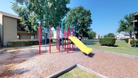 This outdoor playground offers a welcoming recreational space designed for active play and enjoyment. The centerpiece of the area is a multi-element play structure composed of bold red and blue metal poles, supporting a variety of climbing components, monkey bars, a spiral pole, and a bright yellow slide that serves as a standout visual feature. The equipment is thoughtfully arranged to encourage physical activity and exploration for children of varying ages and abilities. The ground beneath the play structure is covered with a generous layer of wood chip mulch, contained by wooden timber borders, providing a safe and cushioned surface. The playground is framed by a lush canopy of mature, leafy trees that offer natural shade and a cool, inviting atmosphere on sunny days. The surrounding grounds are neatly maintained with well-kept green grass, enhancing the open and spacious feel of the area. Residential buildings are visible in the background, suggesting the playground is part of a broader community setting. The clear blue sky above adds to the bright, cheerful ambiance of this well-equipped outdoor space, making it an appealing destination for recreational use.