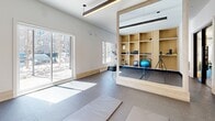 Located at 20 Rue De La Bonne Renomée in Gatineau, Quebec, this single-floor fitness facility offers a thoughtfully designed exercise space built to support a wide range of workout activities. The gym is bathed in natural light courtesy of large windows and a sliding glass door that overlook the surrounding outdoor environment, creating a bright and energizing atmosphere throughout.

The exercise room is divided into two functional zones. The main area is outfitted with a comprehensive selection of fitness equipment, including a multi-station cable machine, elliptical trainers, treadmills, a rowing machine, and a dumbbell rack with an adjustable weight bench — all arranged on durable rubberized flooring. Full-length mirrors line one wall, visually expanding the space while allowing users to monitor their form during workouts. Linear pendant lighting runs along the ceiling, complementing the clean, modern aesthetic defined by white walls and structural columns.

A dedicated functional training area features open rubberized flooring ideal for stretching and mat exercises, with exercise mats readily available. A floor-to-ceiling built-in shelving unit crafted in warm wood tones organizes a variety of smaller equipment, including stability balls, medicine balls, resistance bands, and handheld weights. A wall-mounted television and additional storage cabinetry complete the space, making this facility both practical and visually refined.