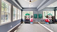 This well-appointed fitness studio at Park View on Denali Drive in Mountain Park, Georgia, offers a bright and functional space designed to support a variety of workout routines. The room features cool-toned, slate-blue walls accented by crisp white trim and baseboards, creating a clean and energizing atmosphere. Textured, low-pile carpet covers the floor throughout, providing a comfortable surface for exercise activities. A large ceiling fan with a wood-beam accent is centrally mounted, ensuring steady air circulation during workouts. Recessed lighting complements the abundant natural light that pours in through multiple large, white-framed double-hung windows, which frame lush greenery and views of an outdoor pool and community area beyond. A full-length wall mirror on one side of the room visually expands the space and supports proper form during exercise. Equipment includes a multi-tier rack stocked with kettlebells, medicine balls, and stability balls of varying sizes, as well as a BOSU balance trainer. Posted fitness center rules and branded signage add an organized, community-oriented touch. French doors with glass panels open to an adjacent interior space, enhancing connectivity and flow. The room is also equipped with a wall-mounted flat-screen display, adding a modern, functional element to the studio environment.