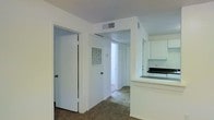 This single-floor residence offers a straightforward and functional layout designed for comfortable everyday living. The interior features neutral white walls and standard-height ceilings throughout, creating a clean and cohesive aesthetic. A hallway serves as the central artery of the space, connecting the various rooms and providing access to what appears to be a bright rear area, allowing natural light to filter through. The kitchen is positioned along one side of the main corridor, equipped with white cabinetry, a dark countertop breakfast bar, and a range, offering a practical cooking and dining setup. Carpeted flooring runs through the main living areas, adding a layer of warmth underfoot. Multiple doorways along the hallway lead to additional rooms, which include closet and utility access, contributing to the overall storage and organizational functionality of the space. A wall-mounted ventilation grille and ceiling HVAC vent are visible, indicating a climate control system integrated throughout. The overall ambiance is modest and utilitarian, presenting a well-organized single-level layout that prioritizes practicality and ease of movement between spaces.