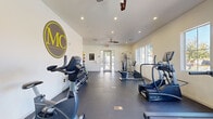This fitness center in Peoria, Arizona offers a dedicated exercise room designed to support a comprehensive workout experience. The space features light-colored walls and durable rubber flooring, creating a clean and functional environment suited for regular physical activity. Ceiling fans and recessed lighting throughout ensure proper air circulation and visibility during workouts.

The room is well-stocked with TRUE-brand cardio equipment, including multiple treadmills, elliptical trainers, a stationary bike, and a rowing machine, providing a wide range of cardiovascular training options. A multi-station cable machine supports strength and resistance training, while a full dumbbell rack with a graduated set of free weights caters to various strength levels. Additional accessories such as resistance bands, medicine balls, foam rollers, and balance balls are also available, offering versatility for functional and flexibility training.

Natural light enters through multiple windows along the walls, complemented by glass-panel entry doors that provide views to the outdoors. A posted fitness rules board helps maintain an organized and respectful environment. The MC branding logo is prominently displayed on the wall, reinforcing the identity of the facility. Overall, the space is thoughtfully arranged to maximize usability while maintaining an open, airy feel.