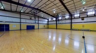 This indoor gymnasium is a well-equipped athletic facility centered around a full-size basketball court finished in polished hardwood. The court is clearly marked with regulation lines in blue and yellow, accommodating multiple game configurations. Multiple basketball hoops are mounted on adjustable backboard systems positioned along the court's perimeter, allowing for versatile play setups.

The facility features a striking high-arched ceiling supported by an exposed steel beam and purlin structural framework, giving the space an expansive, open feel. Overhead, a grid of industrial pendant lights provides consistent, even illumination throughout the court. A row of horizontal windows near the roofline allows natural light to supplement the artificial lighting.

The walls are painted in bold yellow and blue — consistent with West Virginia University's school colors — with a blue wainscoting border along the lower portion. Blue padded wall protectors are installed at key impact zones for player safety. Portable aluminum bleachers are positioned along one sideline, providing seating for spectators.

A set of blue double doors serves as the main entry point, above which a West Virginia University banner is prominently displayed. A first aid station is mounted on the wall nearby. The overall design and branding reflect a strong institutional athletic identity, making this gymnasium well-suited for organized sports, community recreation, and school-level competitions.
