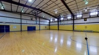 This indoor gymnasium is a well-equipped athletic facility centered around a full-size basketball court finished in polished hardwood. The court is clearly marked with regulation lines in blue and yellow, accommodating multiple game configurations. Multiple basketball hoops are mounted on adjustable backboard systems positioned along the court's perimeter, allowing for versatile play setups.

The facility features a striking high-arched ceiling supported by an exposed steel beam and purlin structural framework, giving the space an expansive, open feel. Overhead, a grid of industrial pendant lights provides consistent, even illumination throughout the court. A row of horizontal windows near the roofline allows natural light to supplement the artificial lighting.

The walls are painted in bold yellow and blue — consistent with West Virginia University's school colors — with a blue wainscoting border along the lower portion. Blue padded wall protectors are installed at key impact zones for player safety. Portable aluminum bleachers are positioned along one sideline, providing seating for spectators.

A set of blue double doors serves as the main entry point, above which a West Virginia University banner is prominently displayed. A first aid station is mounted on the wall nearby. The overall design and branding reflect a strong institutional athletic identity, making this gymnasium well-suited for organized sports, community recreation, and school-level competitions.