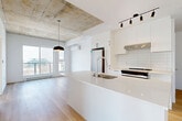Located at 4986 Place De La Savane in Montréal, this contemporary 2-bedroom, 1-bathroom unit on the 8th floor offers a well-designed open-concept layout with a refined industrial aesthetic. The living and kitchen area is the centerpiece of the space, showcasing a striking exposed concrete ceiling that contrasts beautifully with crisp white walls and light hardwood flooring throughout. The kitchen is outfitted with sleek white cabinetry, a subway tile backsplash, stainless steel appliances including a refrigerator and range, and a generous white waterfall island with an undermount sink — ideal for both cooking and entertaining. A wall-mounted air conditioning unit and a large floor-to-ceiling window bathe the living area in natural light while offering open city views. Both bedrooms are bright and airy, featuring the same hardwood flooring, exposed concrete ceiling details, and large windows that welcome ample sunlight. The primary bedroom offers a comfortable footprint with views of the surrounding neighborhood. The bathroom is finished in warm neutral tones, complete with large-format beige tiles, a floating white vanity with integrated sink, a mirrored medicine cabinet, and a full soaking tub with an overhead rain shower. A hallway and dedicated closet add to the unit's practical layout.