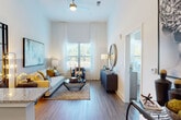 This single-floor residence showcases a contemporary design highlighted by notably tall ceilings that lend an airy, expansive feel to every room. Wood-style flooring runs consistently throughout the space, unifying the open-concept layout and complementing the clean, neutral color palette.

The living area is a welcoming focal point, bathed in natural light from large windows dressed with sheer white curtains and adjustable blinds. A ceiling fan overhead adds both function and style, while the generous proportions comfortably accommodate a full seating arrangement. The space flows seamlessly into the kitchen, which features flat-front cabinetry in a warm wood-tone finish, granite countertops, a tile backsplash, and a full suite of stainless steel appliances including a gas range, refrigerator, built-in microwave, and dishwasher.

The bedroom continues the bright, airy aesthetic with large windows allowing ample natural light and outdoor views. It offers comfortable proportions and direct access to the bathroom, which is equipped with a granite-topped vanity, wood-tone cabinetry, hardwood-style flooring, and a full soaking tub with tiled surround. Throughout, the residence maintains a cohesive modern sensibility with thoughtful finishes and a light, neutral atmosphere.
