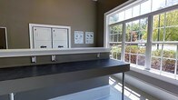 This single-floor communal laundry facility is designed with both functionality and comfort in mind. The room is finished with warm, earthy-toned walls that create a welcoming atmosphere, complemented by crisp white trim and baseboards that lend a clean, polished look throughout.

At the heart of the space are stacked washer and dryer units, neatly arranged along one wall and accompanied by instructional signage for ease of use. A generous dark-surfaced folding counter spans the center of the room, providing ample workspace for residents to sort and fold laundry with ease. The counter features built-in outlets, adding a layer of convenience for users.

One of the standout features of this laundry room is the large, multi-pane window that floods the space with natural light, creating a bright and airy environment. The window frames a lush outdoor view of mature trees and greenery, making the experience of doing laundry far more pleasant than typical utility spaces. The combination of natural light, thoughtful layout, and practical amenities makes this laundry facility a well-considered and highly functional space.