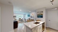 This thoughtfully designed single-floor residence features a bright, open-concept layout that connects the kitchen, dining, and living areas into one cohesive space. The kitchen is outfitted with sleek gray cabinetry, white quartz countertops, stainless steel appliances including a refrigerator, range, built-in microwave, and dishwasher, alongside a peninsula with a stainless steel sink that opens directly to the living and dining area. Light wood-tone flooring runs throughout, lending warmth and visual continuity across all rooms. The living room is generously proportioned, featuring large windows and a glass door that invite ample natural light and offer views of surrounding greenery. Two bathrooms serve the residence: one with a sleek sliding-glass walk-in shower, and a second with a full soaking tub and shower combination, both finished in crisp white with wood-look flooring. The primary bedroom is a comfortable retreat, accommodating a full-sized bedroom set with room to spare, and includes a ceiling fan with lighting, a dedicated closet, and a large window that fills the room with natural light. A hallway and additional closet provide practical storage throughout the home, completing this efficiently designed yet stylish residence.