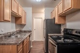 This single-floor, 1-bedroom, 1-bathroom unit offers a practical and efficiently designed layout that makes excellent use of its space. The combined kitchen and living area serves as the heart of the unit, where rich hardwood-style flooring runs throughout. The kitchen is equipped with warm wood cabinetry extending to the upper walls for ample storage, a stainless steel refrigerator, a black electric range, a built-in dishwasher, and a sink with granite-style countertops. A ceiling fan overhead provides airflow into the open living space, which features large windows that allow natural light to fill the room. A dedicated hallway connects the main living area to the private spaces of the unit, including the bedroom, bathroom, and closet. The bedroom is a well-proportioned room featuring plush carpeting, a ceiling fan with light fixture, a baseboard heater, and a window with outdoor views. An attached closet provides convenient storage. The bathroom is clean and functional, featuring wood-style flooring, a vanity with wood cabinetry, a toilet, and a full bathtub with a shower and curtain rod. Neutral wall tones throughout the unit create a calm, cohesive aesthetic.