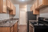 This single-floor, 1-bedroom, 1-bathroom unit offers a practical and efficiently designed layout that makes excellent use of its space. The combined kitchen and living area serves as the heart of the unit, where rich hardwood-style flooring runs throughout. The kitchen is equipped with warm wood cabinetry extending to the upper walls for ample storage, a stainless steel refrigerator, a black electric range, a built-in dishwasher, and a sink with granite-style countertops. A ceiling fan overhead provides airflow into the open living space, which features large windows that allow natural light to fill the room. A dedicated hallway connects the main living area to the private spaces of the unit, including the bedroom, bathroom, and closet. The bedroom is a well-proportioned room featuring plush carpeting, a ceiling fan with light fixture, a baseboard heater, and a window with outdoor views. An attached closet provides convenient storage. The bathroom is clean and functional, featuring wood-style flooring, a vanity with wood cabinetry, a toilet, and a full bathtub with a shower and curtain rod. Neutral wall tones throughout the unit create a calm, cohesive aesthetic.