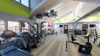 This fitness center is designed to accommodate a wide variety of workout styles within a single, open-plan space. The room's most striking architectural feature is its vaulted ceiling with exposed white-painted wood beams and visible HVAC ductwork, lending an industrial yet polished aesthetic. Clerestory windows positioned along the upper walls allow abundant natural light to fill the space, complementing the recessed track lighting overhead.

The interior is finished with gray walls accented by bold lime-green geometric shapes, creating an energetic and motivating atmosphere. Flooring transitions between rubberized gym flooring in the equipment zones and striped carpet tile in open areas, clearly delineating workout spaces.

Cardio equipment lines one side of the room and includes treadmills and elliptical cross-trainers. The strength training area is well-stocked with multiple racks of dumbbells, adjustable benches, a cable and pulley machine, and TRX suspension trainers. A dedicated functional fitness zone features medicine balls, stability balls, and kettlebells neatly organized on a storage rack. A wall-mounted television is positioned for easy viewing throughout the space. Multiple entry and exit points provide convenient access, with views of an outdoor area visible through ground-level windows.