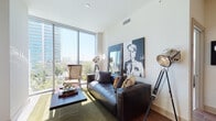 Located at 7901 Windrose Ave in Plano, Texas, this single-floor high-rise residence offers a sleek, contemporary design with an abundance of natural light and striking urban views. The entry hallway sets an elegant tone, featuring wood-look tile flooring and curated wall art that leads seamlessly into the main living areas.

The open-concept kitchen is a standout, boasting a generous island with quartz countertops, pendant lighting, dark wood cabinetry, stainless steel appliances, and a built-in display shelf — all flowing naturally into the dining and living areas. The living room is bathed in sunlight through expansive floor-to-ceiling windows that frame panoramic city skyline views, creating an airy, sophisticated atmosphere.

The primary bedroom is equally impressive, with floor-to-ceiling glass offering sweeping views of the surrounding cityscape and pool area below. A spacious walk-in closet with custom built-in shelving provides generous storage. A secondary bedroom features an elegant tufted headboard wall and natural light from nearby windows.

The bathroom is well-appointed with warm wood vanity cabinetry, a backlit mirror, and a tub-shower combination. Throughout, the residence combines modern finishes, elevated ceilings, and thoughtful design to deliver a refined urban living experience.