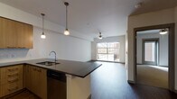 Located in the vibrant Orenco Station neighborhood of Hillsboro, Oregon, this single-floor residence offers a thoughtfully designed open-concept layout that seamlessly connects the kitchen, dining, and living areas. The kitchen is a functional centerpiece, outfitted with warm wood cabinetry, dark countertops, and a full suite of stainless steel appliances including a refrigerator, electric range, built-in microwave, and dishwasher. A peninsula with pendant lighting provides additional workspace and serves as a natural divider between the kitchen and the expansive living area. Rich hardwood-style flooring runs throughout the main living spaces, complemented by neutral walls that create a bright, welcoming ambiance. The living room benefits from a large picture window that frames views of the surrounding neighborhood and fills the space with natural light, while a ceiling fan adds comfort year-round. The bedroom features plush carpeting, its own ceiling fan, and a door providing direct access to an outdoor balcony. The bathroom is clean and functional, featuring a full soaking tub with shower combination and wood-accented vanity. A stacked in-unit washer and dryer add everyday convenience. The overall design balances modern finishes with a warm, livable aesthetic.