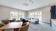 Located at 372 Halstead Ave in Harrison, NY, this single-floor community lounge is a thoughtfully designed gathering space that blends comfort and modern style. The room is finished with soft, neutral-toned walls, recessed lighting, and plush blue-toned carpet throughout, creating a cohesive and inviting atmosphere.

The lounge is divided into distinct functional zones. A relaxed seating area near the expansive floor-to-ceiling French doors features a light blue sofa and coordinating accent chairs in various shades of blue and gray, arranged around low round tables. Multiple sets of French doors flood the space with natural light and open directly onto an outdoor terrace furnished with dining tables and lounge seating, seamlessly extending the space outdoors.

A dedicated dining and collaboration zone includes warm wood-topped tables paired with contemporary rounded upholstered chairs, positioned beneath a wall-mounted flat-screen television — ideal for casual meetings or social gatherings.

The built-in kitchenette is a standout feature, outfitted with bold navy-blue cabinetry, white subway tile backsplash, white countertops, stainless steel appliances including a full-size refrigerator and wall oven, and a coffee station, providing full convenience for residents or guests. A ceiling-mounted HVAC unit ensures year-round climate comfort throughout the space.