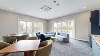 Located at 372 Halstead Ave in Harrison, NY, this single-floor community lounge is a thoughtfully designed gathering space that blends comfort and modern style. The room is finished with soft, neutral-toned walls, recessed lighting, and plush blue-toned carpet throughout, creating a cohesive and inviting atmosphere.

The lounge is divided into distinct functional zones. A relaxed seating area near the expansive floor-to-ceiling French doors features a light blue sofa and coordinating accent chairs in various shades of blue and gray, arranged around low round tables. Multiple sets of French doors flood the space with natural light and open directly onto an outdoor terrace furnished with dining tables and lounge seating, seamlessly extending the space outdoors.

A dedicated dining and collaboration zone includes warm wood-topped tables paired with contemporary rounded upholstered chairs, positioned beneath a wall-mounted flat-screen television — ideal for casual meetings or social gatherings.

The built-in kitchenette is a standout feature, outfitted with bold navy-blue cabinetry, white subway tile backsplash, white countertops, stainless steel appliances including a full-size refrigerator and wall oven, and a coffee station, providing full convenience for residents or guests. A ceiling-mounted HVAC unit ensures year-round climate comfort throughout the space.