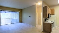 This single-floor residential unit offers a clean, functional layout designed for comfortable everyday living. The main living area is a generous, open space finished with soft wall-to-wall carpeting and neutral warm-toned walls that create a welcoming atmosphere. A full-width sliding glass door fitted with vertical blinds serves as the focal point of the living room, allowing abundant natural light to filter in while providing direct outdoor access and views to the exterior. A wall-mounted thermostat control panel is conveniently positioned within the living space. The textured ceiling adds subtle visual character throughout. Transitioning from the living area, a hallway leads further into the unit, connecting to additional rooms. Adjacent to the living space, the kitchen is accessible and features warm wood-toned cabinetry providing ample storage, along with light-colored countertops. The overall layout flows naturally between the common areas, offering a practical and cohesive living environment. The unit's neutral color palette and consistent flooring throughout contribute to a bright, airy ambiance that is easy to personalize and style.