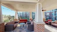 This cabana is a thoughtfully designed outdoor amenity space that seamlessly blends covered comfort with open-air leisure. The structure is anchored by distinctive brick columns with white-painted trim, supporting a covered lounge area fitted with ceiling fans to ensure comfort during warmer days. The covered section features a generous seating arrangement of wicker furniture with striped and solid upholstered cushions, centered around a decorative round coffee table — an ideal setting for casual gatherings or quiet relaxation.

The patio extends beyond the covered area into a fully open-air space, finished with a durable and attractive stone-paver floor throughout. This open section is furnished with high-top tables and chairs, each shaded by bold red market umbrellas, and is strung with overhead bistro lights that lend an inviting ambiance for evening use. The patio offers direct views of a nearby swimming pool, making it a natural extension of the pool area.

Black iron railings define the perimeter of the space, providing both safety and an open sightline to the surrounding landscaped grounds and the multi-story residential buildings beyond. The overall design strikes a balance between functionality and aesthetic appeal, offering multiple seating configurations suited for both lounging and dining in an engaging outdoor environment.