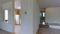 This single-floor space presents a simple, functional layout organized around a central dividing wall that separates the kitchen from the adjoining living areas. The kitchen is visible to one side, equipped with wood-toned cabinetry mounted above the counter space and a full-size white refrigerator. A window in the kitchen allows natural light to filter into the area. The flooring in the kitchen appears to be a light-toned surface, keeping the space bright and easy to maintain. A thermostat is mounted on the central dividing wall, indicating climate control throughout the unit. To the opposite side, the hallway leads toward what appears to be an entry door, with a window nearby that introduces additional natural light into the corridor. The flooring in this section transitions to a darker material, providing a subtle visual distinction between spaces. The overall color palette is neutral, with light-colored walls throughout that contribute to an open and airy feel. The layout is straightforward and efficiently organized, making good use of the available space across the single floor.