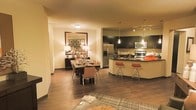 This single-floor residence offers an open-concept layout that seamlessly connects the kitchen, dining, and living areas, creating a cohesive and functional living environment. The kitchen is a standout feature, showcasing dark espresso cabinetry that contrasts elegantly with light-colored walls and countertops. A large stainless steel refrigerator, built-in microwave, and range anchor the cooking space, while a curved kitchen island with a dark countertop provides additional prep space and casual seating with bar-height stools. Pendant lighting above the island adds a refined touch, complemented by a track lighting system overhead. The adjoining dining area is well-defined and inviting, centered around a rectangular dining table with upholstered chairs. A large framed mirror and a classic buffet table with table lamps lend an elegant, warm ambiance to the space. Rich hardwood flooring flows throughout, adding warmth and continuity. Recessed and flush-mount ceiling lighting ensures the entire space is well-illuminated. The overall atmosphere is warm, modern, and thoughtfully designed, balancing contemporary finishes with comfortable, livable touches throughout.