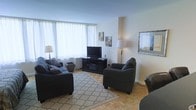 This single-floor space offers a thoughtfully arranged and inviting interior. The living room serves as the centerpiece, showcasing warm parquet hardwood flooring laid in a classic herringbone-style pattern that adds timeless character to the space. Large windows dressed with sheer white curtains allow abundant natural light to fill the room, creating a bright and airy atmosphere throughout the day. The living area is comfortably furnished with a dark upholstered sofa and matching armchairs, anchored by a patterned area rug that defines the seating zone. A sleek, dark media console houses a flat-screen television and serves as a functional focal point along one wall. Tasteful wall art and framed prints add personality and visual interest to the clean, white walls. A classic table lamp provides warm ambient lighting, complementing the natural light. The overall design aesthetic is clean and understated, blending traditional flooring elements with contemporary furnishings for a balanced, livable environment. The layout is well-proportioned, offering a comfortable sense of space without feeling excessive, making it an ideal setting for both relaxing and entertaining.