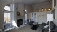 This single-floor residence centers around a striking living room that immediately commands attention with its soaring vaulted ceilings and an impressive whitewashed brick fireplace flanked by tall columns. The fireplace serves as the room's focal point, extending nearly floor to ceiling and complemented by a wide hearth. Large windows, including a distinctive arched window above the fireplace surround, allow generous amounts of natural light to pour in throughout the day, enhancing the open and airy feel of the space. The neutral, warm-toned walls provide a versatile backdrop that complements the room's architectural details. Track lighting along the upper wall adds a modern, adjustable accent to the otherwise classic aesthetic. The living area offers ample room for comfortable seating arrangements, centered around a dark wood coffee table that grounds the space. A frosted glass interior door adds a touch of elegance while maintaining privacy between rooms. The overall ambiance is welcoming and well-balanced, blending traditional architectural character — evident in the brick masonry and arched detailing — with a clean, contemporary sensibility in the furnishings and finishes. The carpeted flooring adds warmth and comfort underfoot, making this a cozy yet visually impressive living environment.