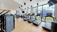 This contemporary fitness center is thoughtfully designed to offer a comprehensive workout experience within a sleek, modern environment. The space is finished with warm light hardwood flooring throughout, which contrasts elegantly against the predominantly white walls and black equipment. Cylindrical surface-mounted light fixtures punctuate the ceiling, providing focused illumination across the floor.

The cardio zone runs along a floor-to-ceiling window wall, where a row of Life Fitness treadmills, ellipticals, and upright bikes face outward toward lush outdoor greenery, flooding the area with natural light. A wall-mounted flat-screen television is positioned nearby for entertainment during workouts, accompanied by a ceiling fan for added airflow comfort.

The strength training area is equally well-appointed, featuring a Synergy 360 functional trainer, adjustable benches, heavy punching bags, and a dedicated free-weight zone with neatly organized dumbbell racks reflected in large floor-leaning mirrors.

A dedicated barre area along one wall includes a mounted ballet barre and wooden stools, ideal for stretching or low-impact training.

A built-in refreshment station with light wood cabinetry, a dark stone countertop, open shelving stocked with towels, and a compact under-counter refrigerator adds a refined, hospitality-driven touch to the overall experience.