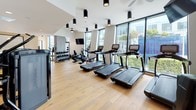 This contemporary fitness center is thoughtfully designed to offer a comprehensive workout experience within a sleek, modern environment. The space is finished with warm light hardwood flooring throughout, which contrasts elegantly against the predominantly white walls and black equipment. Cylindrical surface-mounted light fixtures punctuate the ceiling, providing focused illumination across the floor.

The cardio zone runs along a floor-to-ceiling window wall, where a row of Life Fitness treadmills, ellipticals, and upright bikes face outward toward lush outdoor greenery, flooding the area with natural light. A wall-mounted flat-screen television is positioned nearby for entertainment during workouts, accompanied by a ceiling fan for added airflow comfort.

The strength training area is equally well-appointed, featuring a Synergy 360 functional trainer, adjustable benches, heavy punching bags, and a dedicated free-weight zone with neatly organized dumbbell racks reflected in large floor-leaning mirrors.

A dedicated barre area along one wall includes a mounted ballet barre and wooden stools, ideal for stretching or low-impact training.

A built-in refreshment station with light wood cabinetry, a dark stone countertop, open shelving stocked with towels, and a compact under-counter refrigerator adds a refined, hospitality-driven touch to the overall experience.