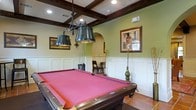 This single-floor space is centered around a well-appointed billiards room that combines classic design with a warm, inviting ambiance. The room's most striking architectural feature is its coffered ceiling, adorned with rich, dark wood beams that create a refined grid pattern overhead. Recessed lighting and an ornate wrought-iron pendant light fixture with dual metal shades hang above the full-size pool table, which is dressed in a bold red felt surface and finished in a deep mahogany wood surround. The walls feature traditional wainscoting along the lower half, painted in crisp white, while the upper portion is finished in a soft sage green tone that adds warmth to the space. Framed artwork decorates the walls, contributing to the curated, sophisticated atmosphere. A graceful arched doorway provides a classic architectural transition to an adjoining area, where seating is available for onlookers and guests. Cue racks are positioned conveniently around the room for easy access. Terracotta tile flooring grounds the space with a timeless, earthy quality. Overall, this space exudes a relaxed yet elegant character, ideal for social gatherings and recreational use.
