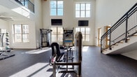 This two-story fitness facility is designed to accommodate a comprehensive range of workout activities across distinct, well-appointed zones. The main floor centers around a dramatic double-height workout area, where towering windows on two levels flood the space with abundant natural light, casting bright patterns across the rubberized flooring. Dual wall-mounted flat-screen TVs and a "Training Zone" equipment cart anchor the open floor plan, which features a variety of strength machines and a cable system. A dedicated strength room off the main area provides a more focused environment with a Smith machine, a weight bench, and rubber flooring for added safety. Full-length mirrors line one wall of the main floor, enhancing the sense of space and supporting proper form during workouts. A decorative staircase with mosaic-style tile risers and wood treads leads to the mezzanine level, where treadmills, a recumbent bike, and additional cardio machines overlook the main floor through an open metal railing. A built-in wood cubby shelving unit on the upper level offers convenient storage. Separate from the main gym, a bright studio room features light hardwood floors, a wall of full-length mirrors, a ceiling fan, recessed lighting, and a wall-mounted TV — ideal for yoga, stretching, or group fitness classes.