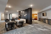 This expansive single-floor amenity lounge is thoughtfully designed to accommodate a variety of social and recreational activities. The space is anchored by striking dark-brick columns that visually define each zone while maintaining an open, connected layout throughout. Patterned carpeting in muted tones of gray, blue, and terracotta flows across the floor, adding texture and warmth underfoot.

At the heart of the lounge, multiple seating arrangements feature a curated mix of contemporary furniture, including curved white sofas, leather armchairs, and sectional couches, providing comfortable spots for relaxed conversation or casual gatherings. A dedicated dining area offers plush velvet booth seating with marble-topped tables, ideal for intimate meals or informal meetings.

A standout game area showcases a full-size billiards table beneath soaring double-height ceilings, framed by a dramatic large-scale graffiti-style mural that adds an urban, artistic edge to the space. Additional wall-mounted televisions are positioned throughout for entertainment. Natural light filters in through floor-to-ceiling windows, offering outdoor views and brightening the interior.

A built-in refreshment station features teal cabinetry with brass hardware and a dark countertop, complemented by a mounted flat-screen television. Warm accent lighting and a wood-paneled bar nook with pendant lights complete the sophisticated yet inviting ambiance of this multifunctional communal space.
