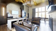 This upscale clubhouse delivers a refined and welcoming atmosphere from the moment you enter. The main living area commands attention with dramatically high ceilings accented by bold gold beam detailing and a striking sunburst chandelier that serves as a luminous centerpiece overhead. Large floor-to-ceiling windows line the perimeter, flooding the interior with abundant natural light and offering open views to the outdoors, while floor-length drapes add an elegant finishing touch.

The open-concept layout seamlessly connects the lounge and social areas. Plush, oversized seating in rich charcoal and taupe tones is arranged to encourage conversation and relaxation. A textured herringbone feature wall frames a built-in media display, adding visual depth and sophistication to the space. Adjacent to the lounge, a stylish wet bar with a white stone countertop and decorative tilework provides an ideal setting for hosting and entertaining.

Underfoot, light-toned flooring in a herringbone pattern complements the warm neutral palette found throughout, while carefully curated décor — including sculptural accents and layered textiles — contributes to the space's polished, contemporary aesthetic. The overall ambiance is both luxurious and inviting, making this clubhouse a standout amenity for community members.