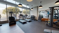 This contemporary fitness center is thoughtfully designed to support a full range of workout routines within a single, open-plan floor. Large floor-to-ceiling windows line the cardio zone, flooding the space with natural light and offering views of the surrounding outdoor streetscape, creating an energizing atmosphere for exercise. Multiple treadmills, a stair climber, and an elliptical machine are positioned along the window wall, complemented by bold black pendant lighting overhead and durable rubber flooring underfoot.

The strength training area features a multi-functional cable/smith machine paired with adjustable benches, framed by full-length mirrors with warm wood-trimmed accents that visually expand the space. A well-organized dumbbell rack holds a comprehensive range of free weights, catering to all fitness levels. An overhead flat-screen television adds entertainment to training sessions.

A dedicated functional training zone transitions to warm hardwood flooring and includes plyo boxes in varying heights for explosive training. Built-in open shelving with warm wood detailing lines one wall, alongside a water refill station, providing a convenient and practical amenity. The overall design balances industrial-modern aesthetics—through concrete-textured walls and matte black fixtures—with inviting warm wood elements, resulting in a polished, motivating environment.