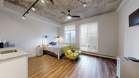 Located on the third floor at 10620 116 Street NW in Edmonton, this guest suite combines industrial-modern design with functional, comfortable living. The exposed concrete ceiling is a defining architectural feature throughout, paired with track lighting and a ceiling fan that together create a dynamic, loft-inspired aesthetic. Hardwood-style flooring runs continuously through the space, adding warmth and cohesion.

The generously sized main room serves as both bedroom and living area. A large window and a sliding glass door fitted with vertical blinds flood the space with natural light and provide direct access to a private balcony with outdoor views. A wall-mounted television and ample open floor space contribute to the room's versatile, comfortable atmosphere.

A compact kitchenette is integrated along one wall, featuring white cabinetry, a single-basin sink, and countertop space accommodating a microwave and coffee maker, making the suite self-sufficient for short-term stays. A connecting hallway leads to the entry door, providing a clear separation between the suite and shared building areas.

Overall, the guest suite offers a well-appointed, self-contained environment with a distinctive industrial character, practical amenities, and good natural light.