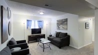 This single-floor space features a well-appointed living area designed for comfort and functionality. The room is finished with plush carpeting underfoot and smooth white walls that create a clean, neutral backdrop. A textured ceiling adds subtle architectural character overhead, complemented by a centrally mounted flush light fixture that provides ambient illumination throughout the space.

The living area is arranged around a charming window dressed with blue patterned curtains, which allows natural light to filter in while adding a touch of color. A dark wood media console sits beneath the window, serving as a practical anchor for the entertainment setup. Opposite the console, a dark gray sofa is paired with a glass-top coffee table featuring a wrought iron base, lending a contemporary yet understated elegance to the room.

Two tall floor lamps flank the window, offering additional warm lighting options. An abstract wall canvas in muted gray and beige tones adds an artistic focal point to the space. The overall ambiance is calm and modern, with a cohesive color palette of dark grays, warm neutrals, and soft whites that makes the space feel inviting and well-balanced.
