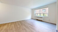 Located on Queen Street East in Toronto's Upper Beaches neighbourhood, this three-bedroom, one-bathroom unit offers a thoughtfully updated interior across a single floor. Light wood laminate flooring runs throughout, complementing the clean white walls and creating a bright, cohesive feel.

The open-concept kitchen and living area forms the heart of the unit. The kitchen is outfitted with white cabinetry, dark stone countertops, a herringbone tile backsplash, and stainless steel appliances including a French door refrigerator, dishwasher, and range. The adjoining living area is spacious and filled with natural light from a large multi-pane window, with a radiator unit positioned below.

The primary bedroom is a well-proportioned room featuring a corner window that invites greenery views and natural light. A closet with panelled doors provides dedicated storage. The two additional bedrooms are similarly bright, each with windows and light-toned walls — one featuring a radiator beneath a window framing lush ivy-covered surroundings.

The bathroom is cleanly finished with large-format grey floor tiles, white subway tile surrounding the soaker tub, and a window for natural ventilation. A hallway connects the rooms efficiently, with a dedicated closet offering additional storage. Elegant three-panel interior doors are featured throughout the unit.