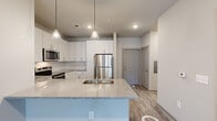 This well-appointed one-bedroom, one-bathroom residence in Suwanee, GA offers a thoughtfully designed open-concept layout with modern finishes throughout. The combined kitchen and living area serves as the heart of the home, featuring durable wood-style plank flooring that extends seamlessly across the entire unit. The kitchen is equipped with a full suite of stainless steel appliances, including a refrigerator, range, microwave, and dishwasher. White shaker-style cabinetry with ample storage reaches to the ceiling, while a large center island with a granite countertop and undermount sink provides additional prep space and a casual dining option. Stylish pendant lighting hangs above the island, complementing the recessed lighting throughout. The open living area benefits from natural light streaming through large windows and a glass-paneled door that leads to a private outdoor area, with a ceiling fan adding comfort year-round. The bedroom is a comfortable, well-proportioned retreat with a ceiling fan, natural light from dual windows, and access to an outdoor balcony. The bathroom features a tub-shower combination with tile surround, a granite-topped vanity, and wood-style flooring. A hallway connects the living spaces, completing this clean, contemporary residence.