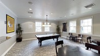 This single-floor recreational space is designed for leisure and social engagement, centered around a standout billiards room that serves as the heart of the layout. The room features an ornate pool table with richly carved dark wood legs and a clean white felt surface, positioned beneath a decorative pendant chandelier with warm-toned glass shades that adds a touch of classic elegance. Recessed lighting throughout the textured ceiling ensures the space is well-lit at all times, while multiple white-framed windows along the walls flood the room with natural light and offer views of the surrounding greenery. Crown molding lines the upper perimeter of the walls, lending the space a polished, refined character. The flooring is covered in a patterned carpet that adds warmth and texture underfoot. A mounted cue rack on one wall keeps equipment neatly organized, while additional seating and table arrangements along the periphery provide comfortable spaces for spectators or casual conversation. Potted plants add a touch of natural warmth to the corners of the room. Overall, the space strikes a balance between functionality and sophisticated design, making it well-suited for both competitive play and relaxed social gatherings.