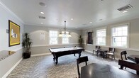 This single-floor recreational space is designed for leisure and social engagement, centered around a standout billiards room that serves as the heart of the layout. The room features an ornate pool table with richly carved dark wood legs and a clean white felt surface, positioned beneath a decorative pendant chandelier with warm-toned glass shades that adds a touch of classic elegance. Recessed lighting throughout the textured ceiling ensures the space is well-lit at all times, while multiple white-framed windows along the walls flood the room with natural light and offer views of the surrounding greenery. Crown molding lines the upper perimeter of the walls, lending the space a polished, refined character. The flooring is covered in a patterned carpet that adds warmth and texture underfoot. A mounted cue rack on one wall keeps equipment neatly organized, while additional seating and table arrangements along the periphery provide comfortable spaces for spectators or casual conversation. Potted plants add a touch of natural warmth to the corners of the room. Overall, the space strikes a balance between functionality and sophisticated design, making it well-suited for both competitive play and relaxed social gatherings.