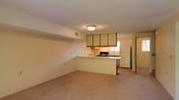 This two-floor space features an open-concept main level where the living area and kitchen flow seamlessly together. The living area is carpeted in neutral tones, providing a warm and understated foundation that complements the overall palette of the space. A flush-mount ceiling light fixture illuminates the room, and a visible HVAC vent is integrated into the upper wall, reflecting practical utility.

The kitchen is defined by a peninsula-style counter that separates it from the living space, offering both a workspace and a natural division between the two areas. Upper cabinetry with wood-trimmed doors provides storage above the countertops, while a built-in range and a full-size refrigerator are positioned along the kitchen's rear wall. A window above the sink allows natural light to filter into the kitchen area, brightening the workspace. The countertops are finished in a dark tone, contrasting with the lighter cabinetry and walls.

The walls throughout are painted in a clean, off-white tone, contributing to a bright and neutral ambiance. A doorway visible near the kitchen hints at additional rooms or access points within the layout, and the second floor adds further living or functional space to the overall unit.