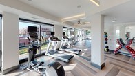 This contemporary fitness center is thoughtfully designed to support a full range of exercise disciplines across a generously sized, single-floor layout. The space is divided into distinct functional zones, each finished to a high standard and bathed in natural light from expansive floor-to-ceiling windows that frame outdoor views on multiple sides.

The cardio zone is lined with treadmills and elliptical machines, each equipped with individual screens for entertainment during workouts. Herringbone-patterned flooring in warm neutral tones adds a refined aesthetic touch throughout this area, complemented by ceiling fans and recessed linear lighting that maintain a bright, comfortable environment.

Adjacent to the cardio area, the strength training zone features a variety of cable and resistance machines, along with free-weight equipment, all arranged to allow ample circulation space. Bold navy cabinetry provides stylish storage solutions along one wall, adding a striking design contrast to the predominantly white and light-toned interior.

A dedicated studio or functional training room offers a more open, flexible space finished with darker hardwood-style flooring. Large windows flood the room with sunlight, while a circular pendant light serves as a focal design element. Resistance bands, medicine balls, and stability equipment are neatly organized on racks, making this area ideal for group classes or individual functional training sessions.