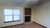 This single-floor space offers a comfortable and functional layout centered around a standout living area. The room's focal point is a tile-surround fireplace accented by a decorative wood chevron panel above the firebox, adding a warm, rustic character to the space. The fireplace is framed in classic square tile extending to a raised hearth, blending a traditional aesthetic with practical design.

Natural light filters into the living area through a large window fitted with horizontal blinds, offering pleasant views of lush, mature trees outside. The soft carpeted flooring throughout contributes to the room's cozy atmosphere.

Overhead, a combination of track lighting and pendant light fixtures provides versatile illumination options, allowing the space to be lit according to function or mood. The ceiling fan adds to the comfort and airflow of the room. Neutral, cream-toned walls keep the space feeling bright and open, providing a clean backdrop that complements the earthy tones of the carpet and fireplace tile.

Overall, this space strikes a balance between functional living and inviting warmth, with its fireplace and natural light serving as key architectural highlights throughout the interior.