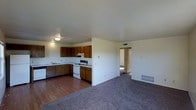 This single-floor residence offers a practical and open layout designed to maximize everyday functionality. The kitchen is outfitted with warm wood-toned cabinetry arranged in an L-shaped configuration, providing ample storage and counter space. It includes a white electric range, a built-in dishwasher, and a sink positioned along the cabinetry. The kitchen features durable hardwood-style flooring, and overhead lighting keeps the workspace well lit. A half-wall partition subtly defines the kitchen from the adjoining living area, which transitions to plush, wall-to-wall carpeting that adds warmth and comfort underfoot. The living area is open and spacious, benefiting from a clean, neutral color palette on the walls and ceilings that enhances the sense of brightness throughout. A wall-mounted heating unit is conveniently located in the living space, ensuring efficient climate control. A hallway visible from the main living area leads to additional rooms, suggesting a thoughtful floor plan that separates the common and private spaces. Overall, the residence presents a straightforward and functional design, well-suited for comfortable single-level living.
