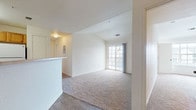 This single-floor residence offers a well-organized layout designed for comfortable everyday living. The open-concept living area features soft carpeting and is anchored by a set of sliding glass doors that lead to a private balcony, allowing natural light to fill the space. Vaulted ceilings in the living room add a sense of height and openness, while neutral wall tones throughout create a clean, cohesive aesthetic.

The kitchen is thoughtfully positioned adjacent to the living area, separated by a half-wall peninsula that functions as a breakfast bar. It is equipped with warm honey-toned wood cabinetry, light countertops, white appliances including an electric range and dishwasher, and a built-in overhead range hood. Wood-look flooring in the kitchen and entry areas provides durability and a visual contrast to the carpeted living spaces.

The bedrooms are comfortably sized, each featuring plush carpeting and windows that frame views of surrounding trees, providing both natural light and a sense of privacy. The bathroom is finished with wood-look vinyl flooring, a full bathtub and shower combination, a vanity with wood cabinetry, and clean white fixtures throughout. Overall, the space presents a functional and well-maintained interior suited for a range of living needs.