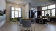 This stylish single-floor space is designed with an open-plan layout that seamlessly connects a fully equipped kitchen to multiple dining and lounge areas. The kitchen is appointed with sleek dark cabinetry, granite countertops, a stainless steel refrigerator, and a functional island with bar seating, making it ideal for casual gatherings and social events. Pendant lighting above the island adds a refined touch, complementing the recessed ceiling lights throughout the space. Multiple dining arrangements cater to different group sizes — a high-top bistro table with contemporary chrome-based chairs sits at the center, while a secondary dining area with round tables and vibrant accent seating is visible near the large windows. The light hardwood flooring and neutral wall tones create a clean, welcoming ambiance, while soaring ceiling heights enhance the sense of openness. Generous windows along the perimeter flood the interior with natural light and offer views of the surrounding greenery. The overall aesthetic is modern and sophisticated, with a cohesive palette of grey, white, and dark tones that give the space a polished, communal character.